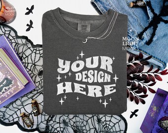 May include: Dark gray t-shirt with the text "YOUR DESIGN HERE" in white, surrounded by stars. The shirt is part of a Halloween-themed flat lay with books, a candle, and decorative elements.