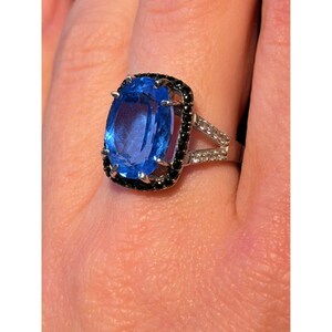 Könnte beinhalten: Ein silberfarbener Ring mit einem großen, oval geschliffenen blauen Edelstein als Mittelpunkt. Der blaue Stein ist von einem Kranz aus kleinen, schwarzen Steinen umgeben. Die Ringschiene ist geteilt und mit kleinen, klaren, weißen Steinen besetzt.