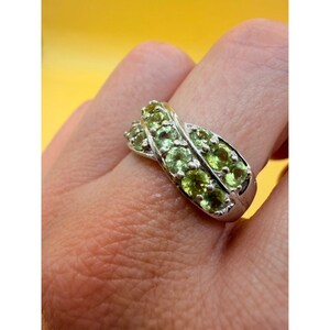 Könnte beinhalten: Ein silberfarbener Ring mit einem überkreuzten Design, besetzt mit mehreren runden, hellgrünen facettierten Steinen. Die Ringschiene hat eine polierte metallische Oberfläche und die Steine sind in zwei parallelen Reihen angeordnet.