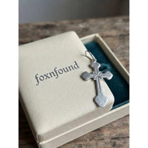 Peut inclure: Un pendentif en forme de croix en métal argenté avec une gravure texturée en forme d'étoile au centre et une petite bélière circulaire au sommet. Le pendentif repose sur un couvercle de boîte crème avec le mot foxnfound imprimé en gris foncé.