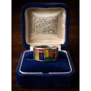May include: A silver-toned band ring featuring a mosaic of rectangular translucent enamel inlays in red, orange, yellow, green, and blue. The ring is displayed inside a dark blue velvet jewellery box with the text foxnfound and a fox illustration.