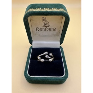 May include: A silver-toned ring with a multi-layered, open-ended design featuring rounded, ball-like terminals at each end. The ring is displayed inside a dark green velvet jewelry box with a cream-colored interior featuring a fox logo and the text foxnfound.