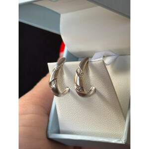 May include: A pair of silver-toned hoop earrings featuring a twisted, ribbed design with small, clear embedded stones along the center. The earrings are displayed on a white textured insert inside a light blue jewelry box.