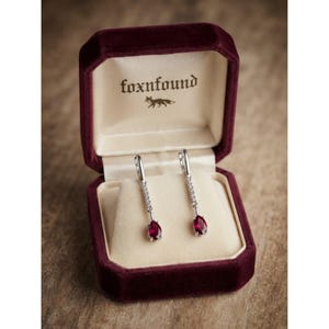 May include: A pair of silver drop earrings featuring a vertical row of small clear stones and a single pear-shaped deep red gemstone at the base. The earrings are displayed inside a dark burgundy velvet jewellery box with the text foxnfound.