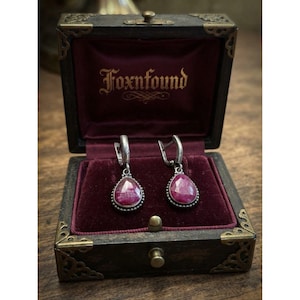 May include: A pair of silver drop earrings featuring faceted, pear-shaped red stones, displayed inside a dark wooden box with a deep red velvet lining. The interior lid of the box features the gold-colored text Foxnfound in a script font.