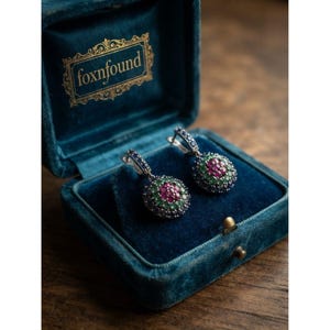 Puede incluir: Un par de pendientes redondos con un grupo central de piedras rosas rodeado por un anillo de piedras verdes y un borde exterior de piedras azul oscuro, presentados en un joyero de terciopelo azul verdoso con el texto dorado foxnfound.
