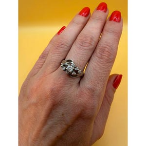 May include: A silver-toned ring featuring a central round-cut clear stone surrounded by a cluster of smaller clear stones. The band has a bypass-style design that curves around the central setting, creating a layered appearance on the finger.