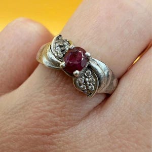Peut inclure: Bague en argent ornée d'une pierre rouge foncé ronde au centre, flanquée de petits brillants transparents. La bague présente un motif tourbillonnant décoratif sur le corps.