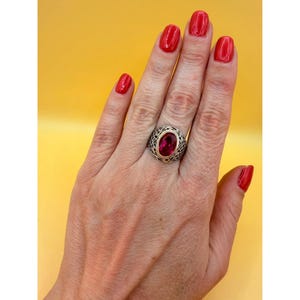 May include: A silver ring featuring an oval-shaped, deep pink gemstone set in a gold-toned bezel. The band of the ring displays an intricate, openwork lattice pattern that extends around the finger, providing a detailed and textured appearance.