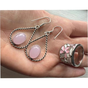 May include: A pair of silver-toned drop earrings featuring twisted metal frames and oval-shaped light pink stones, accompanied by a wide silver-toned ring decorated with white and pink enamel floral patterns and small clear inset stones.