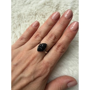 May include: A silver-toned ring featuring a marquise-shaped centerpiece encrusted with small, dark black crystals. The setting is metallic and polished, showcasing a textured, faceted surface on the central stone element that catches the light.