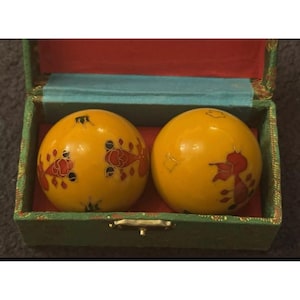May include: Two yellow spherical balls featuring red and black koi fish illustrations, housed in a rectangular green fabric-covered box with a red interior lining and a light blue fabric trim along the inner top edge of the container.