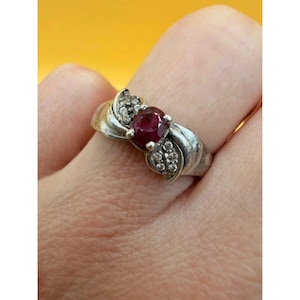 May include: A silver-toned ring featuring a central oval-cut deep red gemstone. The band is accented with small, clear, round-cut stones embedded in a textured, swirling metal design that frames the central red stone on both sides.