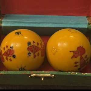 Puede incluir: Dos bolas decorativas amarillas con diseños florales rojos y negros se exhiben dentro de una caja verde con interior turquesa. La caja tiene un cierre dorado y un forro de terciopelo rojo.