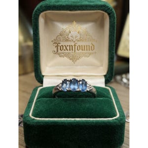 May include: A silver-toned ring featuring three oval-cut blue stones and small clear accent stones on the band. The ring is displayed inside a dark green velvet jewellery box with the gold-coloured text Foxnfound and a fox logo printed inside.