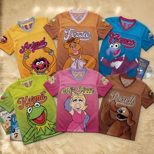 May include: Six V-neck jerseys featuring Muppets characters. The yellow shirt shows Animal, the tan shirt shows Fozzie, the blue shirt shows Gonzo, the green shirt shows Kermit, the pink shirt shows Miss Piggy, and the brown shirt shows Rowlf.
