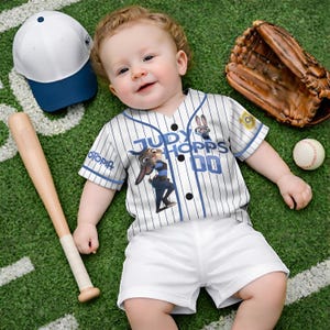 Puede incluir: Un beb&eacute; con un uniforme de b&eacute;isbol con una camiseta "Judy Hopps 00", pantalones cortos blancos y una gorra de b&eacute;isbol. Un bate, un guante y una pelota de b&eacute;isbol est&aacute;n en un campo verde.