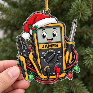 May include: A clear acrylic ornament featuring a cartoon multimeter wearing a red Santa hat with holly. The multimeter has a yellow body, a screen displaying the name JAMES, a dial, and the year 2026. It is surrounded by wire strippers, a screwdriver, and colorful Christmas lights.