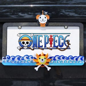 Puede incluir: Matrícula de coche con el texto "ONE PIECE" y personajes de dibujos animados. El marco tiene un diseño de olas azules y blancas en la parte inferior. Un perro de dibujos animados y un sol con huesos cruzados están adjuntos.