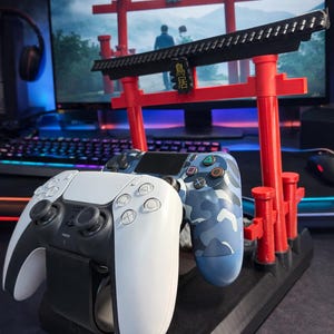 May include: A black and white PlayStation 5 controller rests on a black charging stand, next to a blue camouflage controller. A red and black Japanese Torii gate decorates the stand. The background shows a computer monitor with a landscape scene.