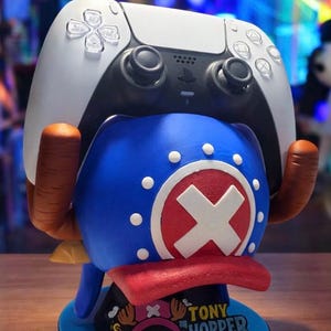 May include: A blue, red, and white Tony Chopper-themed controller stand holding a white and black PlayStation 5 controller. The stand features a Chopper design with a red cross and the text "TONY CHOPPER" on a blue base.