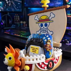 May include: A One Piece Thousand Sunny ship-shaped charging station for a game controller. The ship features a yellow and orange lion figurehead, a white sail with a skull and crossbones, and a black flag. The controller has a blue design with a character illustration.