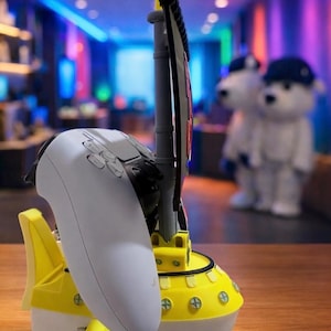 May include: A yellow and white gaming controller charging station shaped like a ship. The station has a mast and sail design, with a white controller resting in the cradle. The background is blurred, showing a room with colorful lights.