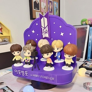 Könnte beinhalten: Ein lila Display mit sieben Tiny Tan Figuren. Das Display hat eine geschwungene Oberseite mit Sternaussparungen und dem Tiny Tan Logo. Auf der Basis steht "Army Forever" in weißem Text. Die Figuren haben unterschiedliche Haarfarben und Outfits.