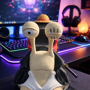 May include: A detailed figurine of a snail-like character with large, expressive eyes and a straw hat. The figure is wearing a black coat and a white shirt. A small sword is attached to the side. The background includes a computer monitor and keyboard.