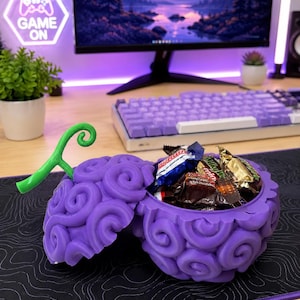 May include: A purple, brain-shaped container, open to reveal wrapped candies. The container has a green stem and sits on a black mousepad with a white topographic design. A computer monitor and keyboard are in the background, with a "Game On" neon sign.