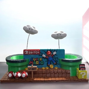 May include: A Super Mario-themed display featuring a brick platform, green pipes, and a blue backdrop with the Nintendo Switch logo. Mario is depicted breaking through the backdrop. Includes cloud decorations, gold coins, and a "Start" sign.