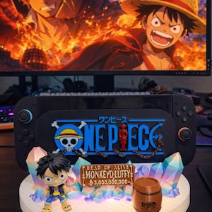 May include: A black Nintendo Switch console with a One Piece theme, featuring a figure of Monkey D. Luffy. The base has a sign that reads "DEAD OR ALIVE MONKEY D. LUFFY 3,000,000,000." A monitor in the background displays One Piece characters.