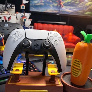 May include: A white and black PlayStation 5 controller on a brown stand with the text "TO CARROTS From Your Emotional Support Animal". A carrot-shaped item sits in a pot. A pirate ship model is in the background.