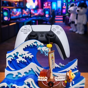 May include: A white and black PlayStation 5 controller rests above a decorative wave and ship scene. The wave is blue and white, and the ship is brown with a yellow sail. The scene is set against a gaming room background.