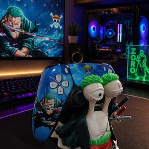 May include: A blue and black video game controller featuring a character from the anime series One Piece. A figurine of a snail with swords and a green wig is in front of the controller. A computer monitor and keyboard are in the background.