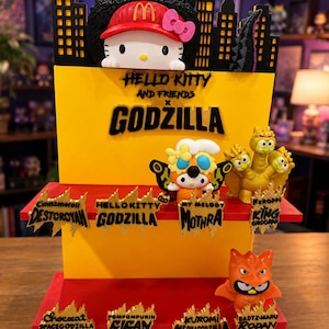 Op de afbeelding: Een displaystandaard met meerdere niveaus voorzien van Hello Kitty and Friends x Godzilla branding. Bovenaan staat een Hello Kitty figuur met een McDonald's pet en Godzilla erachter. Lagere niveaus bevatten karakterfiguren en gouden naamplaatjes.