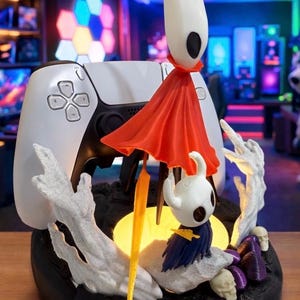 May include: A detailed figurine featuring characters from the video game Hollow Knight, with a white controller in the background. The figurine includes a character with a red cape and another with a blue cape, set on a black base with a glowing yellow light.