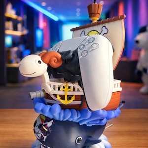 May include: A white PlayStation 5 controller is integrated into a whimsical pirate ship design. The ship features a brown hull, a white sail with a skull and crossbones, and a black flag. The base is a blue whale with a skull and crossbones design.