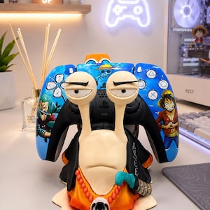 May include: A custom video game controller stand shaped like a One Piece snail character. The stand features a blue controller with anime character graphics, while the snail base is painted in orange and black with a skull pendant and bead necklace.