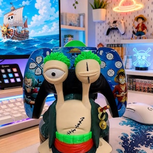 Może przedstawiać: Niestandardowy kontroler do gier wideo z postacią z anime One Piece. Kontroler ma zielono-czerwoną kolorystykę, z twarzą i detalami postaci. Kontroler jest umieszczony przed monitorem komputera.