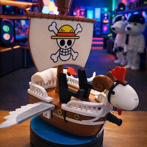 May include: A detailed model of the Thousand Sunny ship from the One Piece anime. The ship features a white sail with a skull and crossbones, a brown hull, and a ram head figurehead. It sits on a blue base.