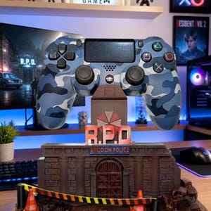 以下が含まれることがあります： 青い迷彩柄のPlayStation 4コントローラーが、詳細なラクーン市警（R.P.D.）のディスプレイの上に置かれています。ディスプレイには、R.P.D.のロゴと注意テープのバリアが付いたレンガ造りの建物があります。「EAT SLEEP GAME」という文字が見えます。