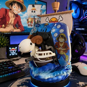 May include: A blue and white video game controller with a One Piece theme, featuring Luffy. The controller is mounted on a ship-shaped stand with a sheep figurehead and a blue wave base. A monitor in the background displays a One Piece scene.
