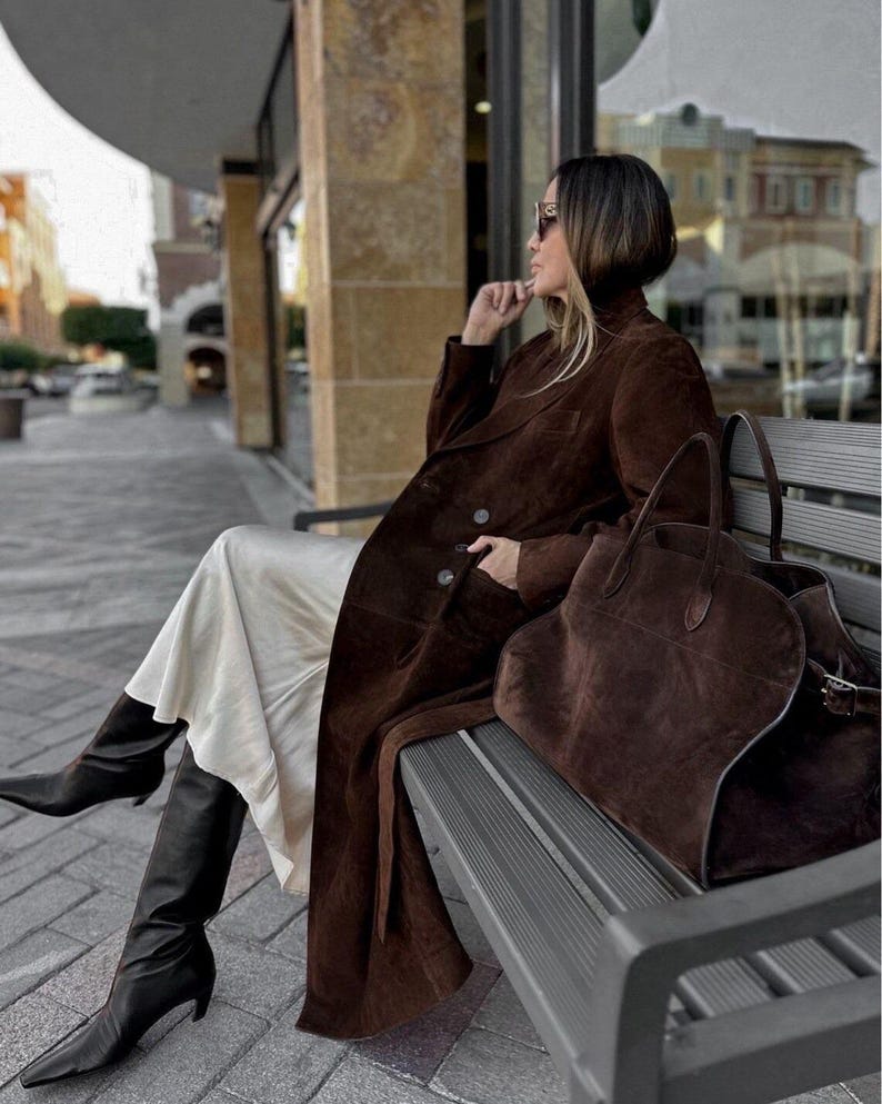 Peut inclure: Une personne porte un long manteau en daim marron, des bottes noires montantes et des lunettes de soleil, assise sur un banc. Un grand sac fourre-tout en daim marron est pos&eacute; &agrave; c&ocirc;t&eacute;. Le manteau est crois&eacute;.