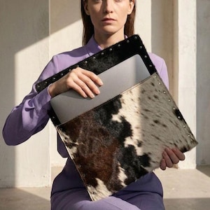 Può includere: Una custodia per laptop in pelle di mucca con un motivo maculato bianco e nero e finiture in pelle nera. La custodia è tenuta da una persona che indossa una camicia viola.