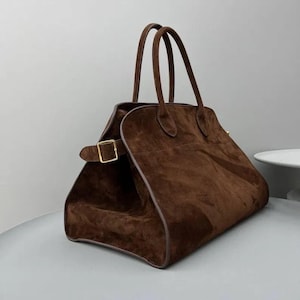 Peut inclure: Sac &agrave; main en daim marron avec deux anses et une forme rectangulaire. Le sac est dot&eacute; d'une fermeture &agrave; boucle dor&eacute;e et d'un design structur&eacute;. Le sac est d'un riche brun fonc&eacute;.