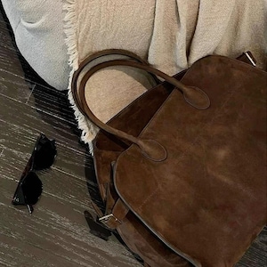 Peut inclure: Un sac fourre-tout en daim marron avec deux poign&eacute;es et une fermeture &agrave; boucle. Le sac est ouvert, r&eacute;v&eacute;lant l'int&eacute;rieur. Des lunettes de soleil noires reposent &agrave; c&ocirc;t&eacute; du sac sur une surface en bois fonc&eacute;. Le sac est de couleur neutre.