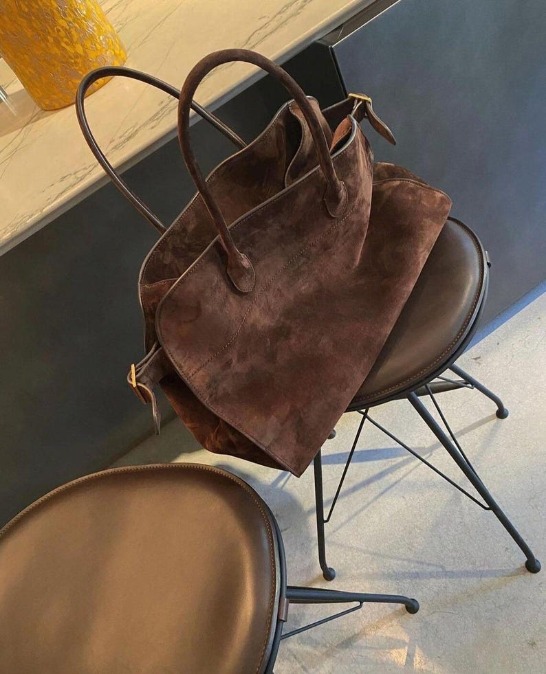 Peut inclure: Un sac fourre-tout en daim marron avec deux poign&eacute;es repose sur une chaise en cuir marron avec des pieds en m&eacute;tal noir. Le sac a une forme rectangulaire et un aspect doux et textur&eacute;. La chaise est ronde avec une assise marron fonc&eacute;.