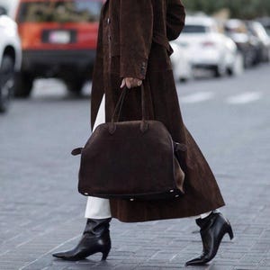 Peut inclure: Une personne marchant dans la rue, v&ecirc;tue d'un long manteau marron, d'un pantalon blanc et de bottes noires. Elle porte un grand sac &agrave; main marron fonc&eacute; avec une poign&eacute;e sup&eacute;rieure. L'arri&egrave;re-plan comprend des voitures.
