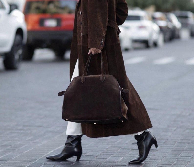 Peut inclure: Une personne marchant dans la rue, v&ecirc;tue d'un long manteau marron, d'un pantalon blanc et de bottes noires. Elle porte un grand sac &agrave; main marron fonc&eacute; avec une poign&eacute;e sup&eacute;rieure. L'arri&egrave;re-plan comprend des voitures.